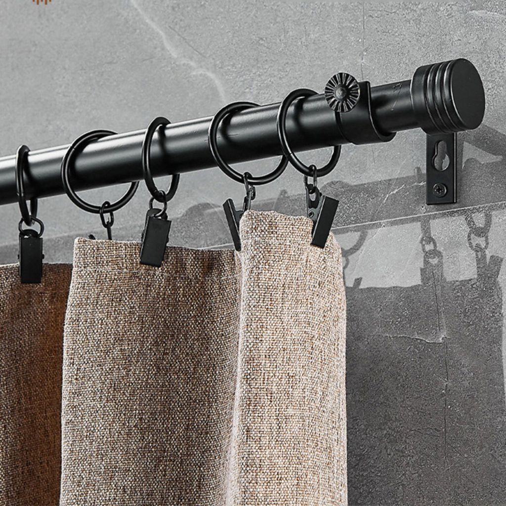 A close-up view of a black curtain rod with rings and clips holding up beige curtains against a gray tiled wall.