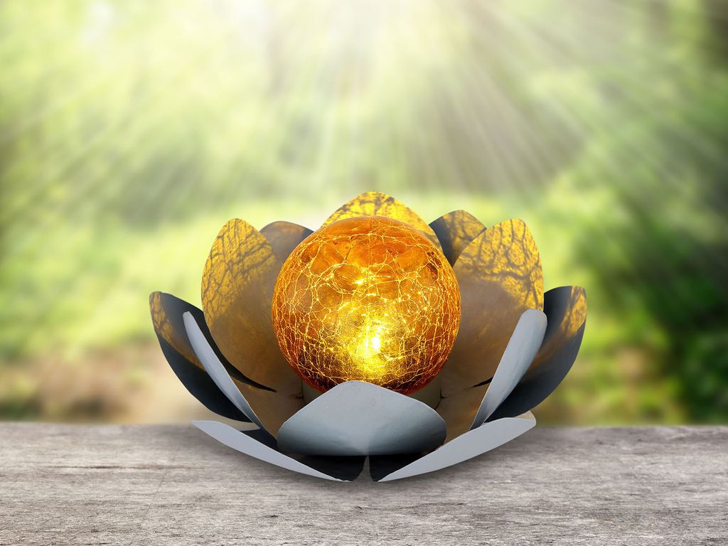 A decorative solar light shaped like a lotus flower with a glowing orb inside, placed on a wooden surface with a blurred green background of trees and sunlight.