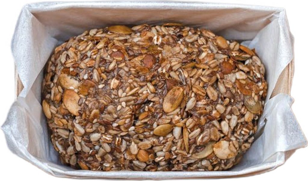 A loaf of bread covered in seeds and grains, placed inside a baking pan lined with parchment paper.