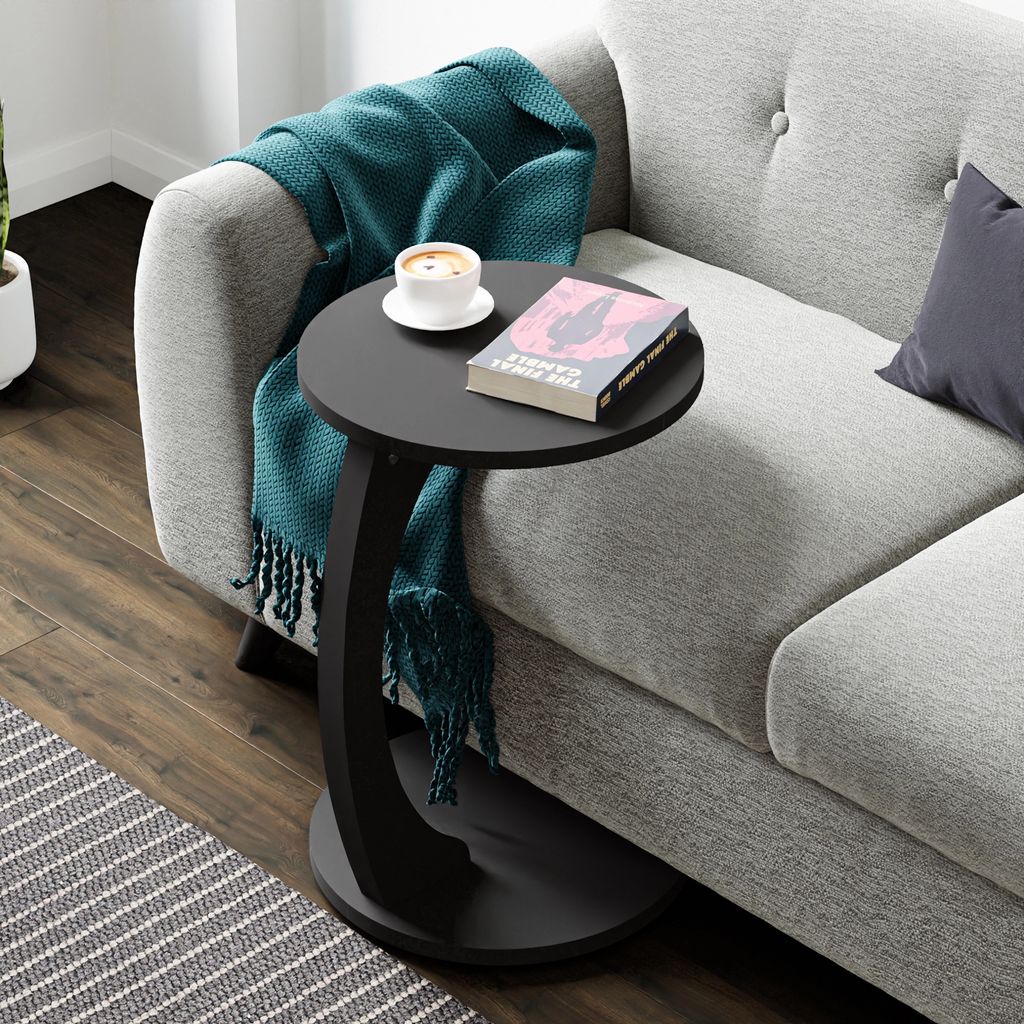 A black C-shaped side table with a book and a cup of coffee on it, placed next to a gray sofa with a teal throw blanket.