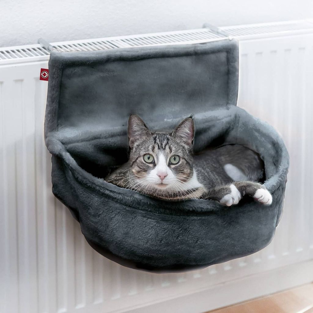 A cat is resting in a gray fabric hammock attached to a white radiator.