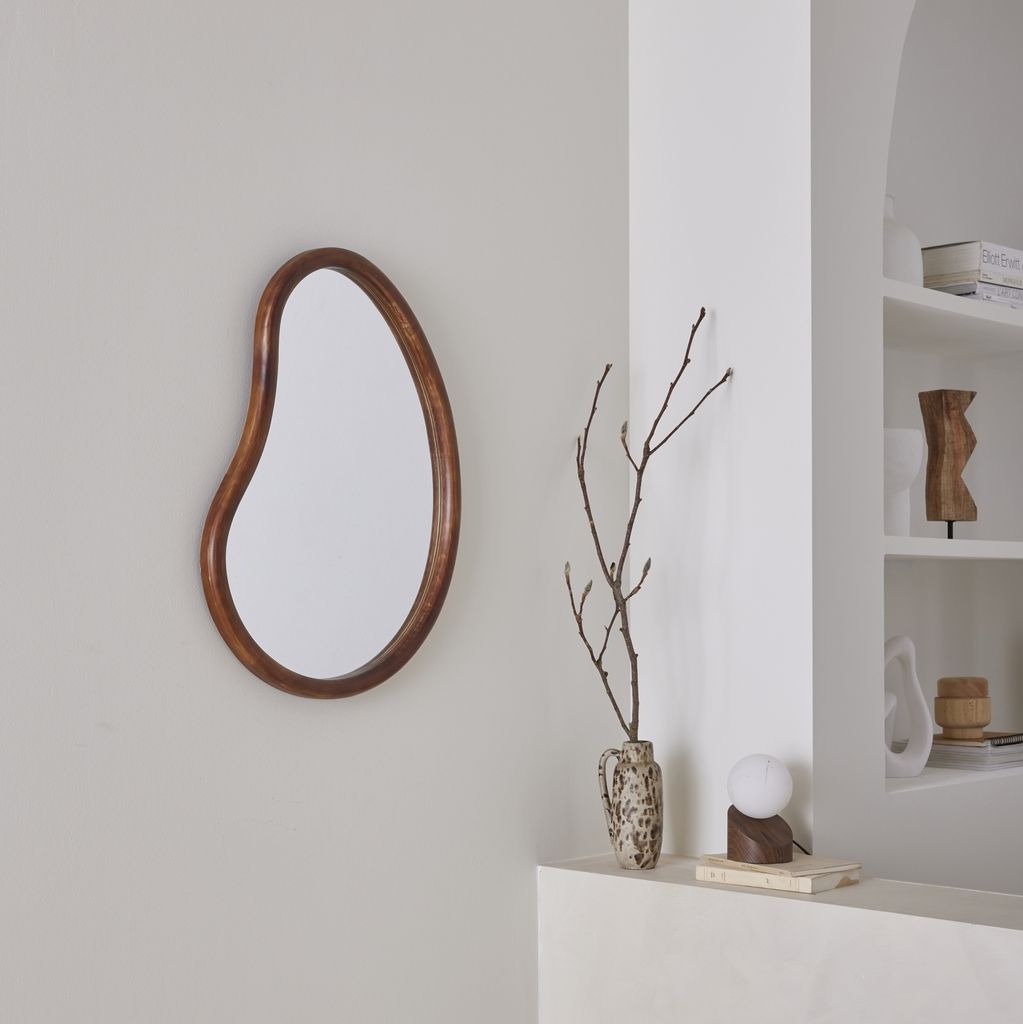 A modern living room corner with a mirror, vase with branches, and a lamp on a shelf.