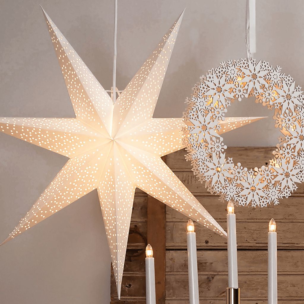 A large, white, star-shaped paper lantern with small perforations hangs next to a white snowflake wreath. Several white LED candles are arranged in a holder in the foreground.
