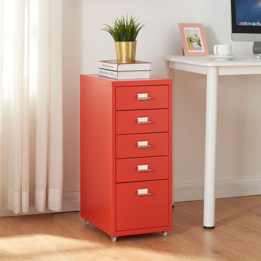 A red filing cabinet with five drawers is placed next to a white desk. There are books and a plant on top of the cabinet. A computer monitor and a cup of coffee are on the desk.