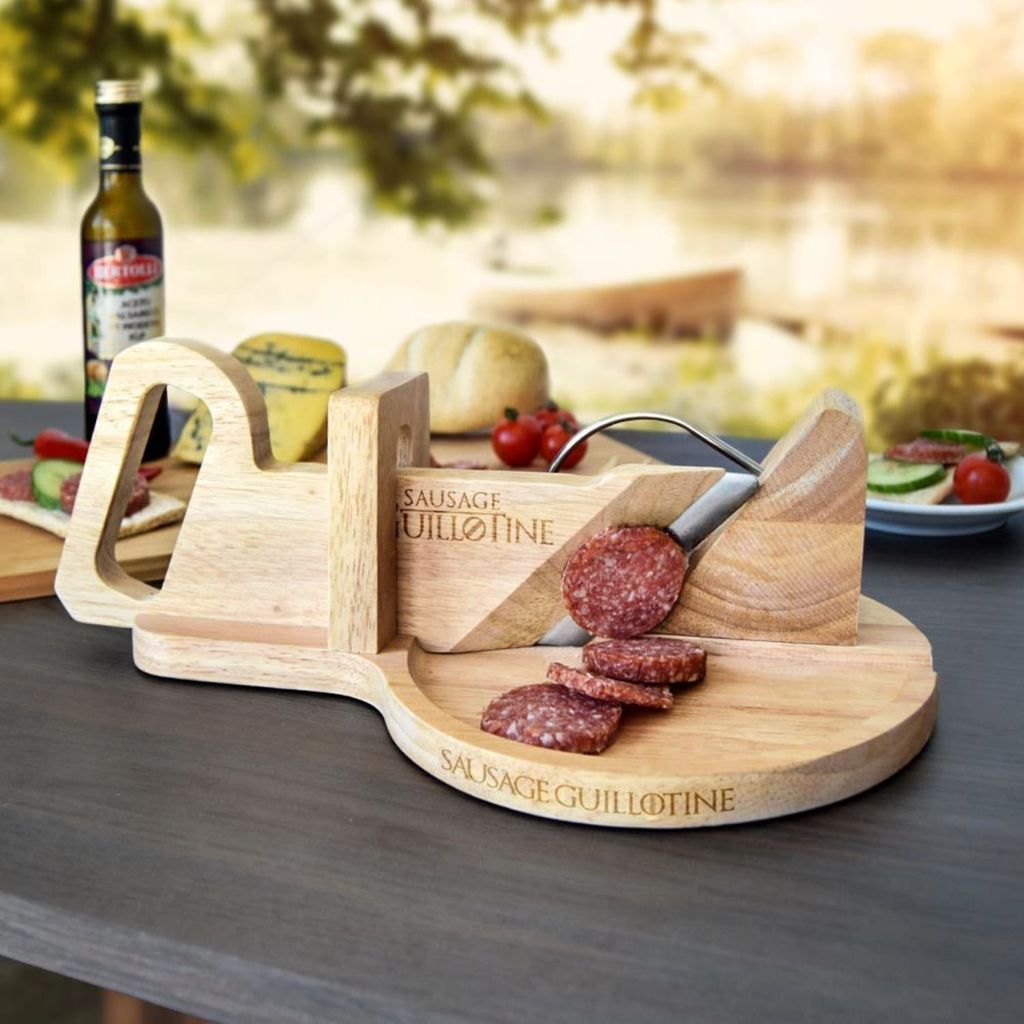A wooden sausage guillotine is set up outdoors on a table, with food items like cheese, bread, and tomatoes in the background, suggesting a picnic or outdoor meal setting.