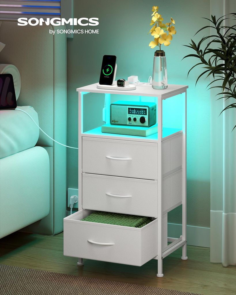 A white nightstand with drawers and an open shelf, featuring a phone charging, a radio, and a vase with flowers. The nightstand has integrated LED lighting.