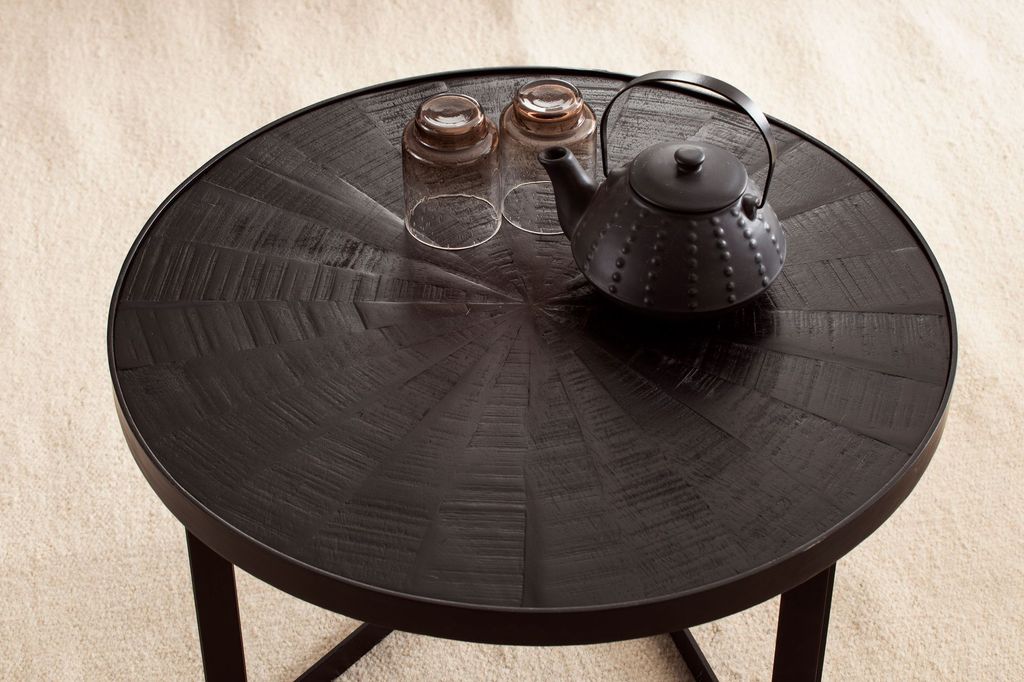 A round black coffee table with a textured surface, two glasses, and a black teapot on top, placed on a light beige carpet.