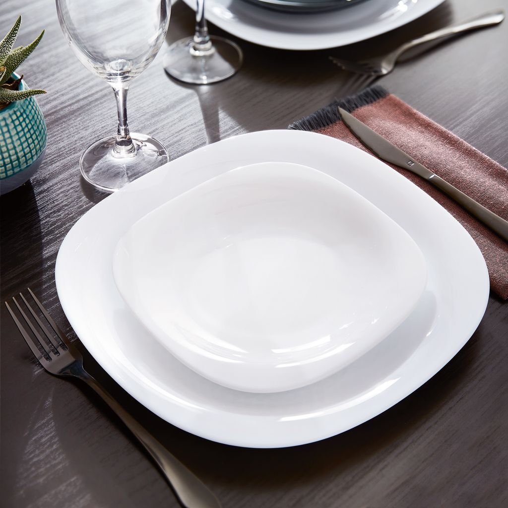 A table setting with white square plates and a bowl, a wine glass, and cutlery.