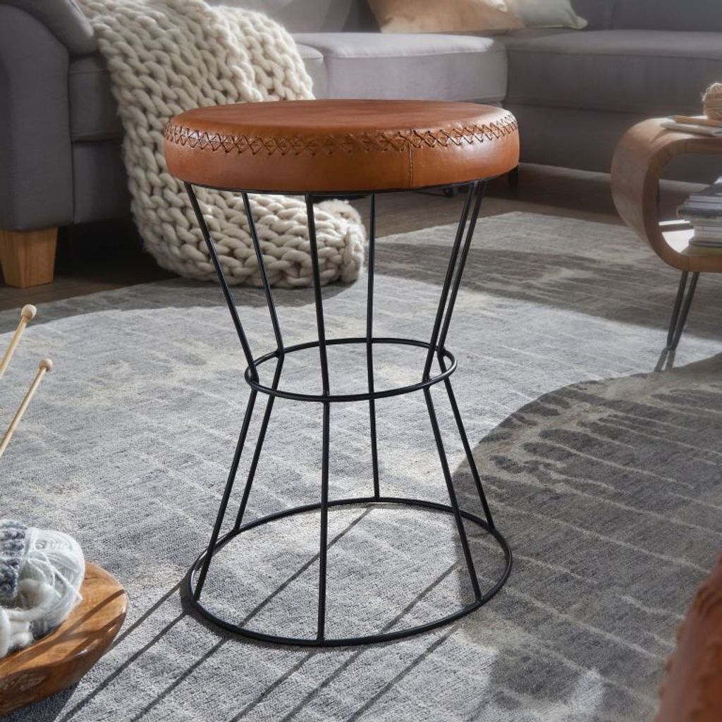 A brown leather stool with a black wire base sits on a gray rug in a living room setting.