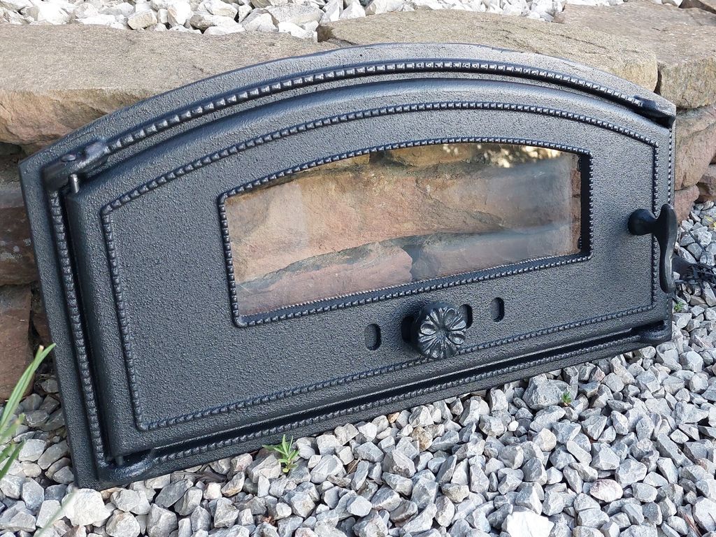 A black cast iron oven door with a glass window rests on a bed of gray gravel and large stones.