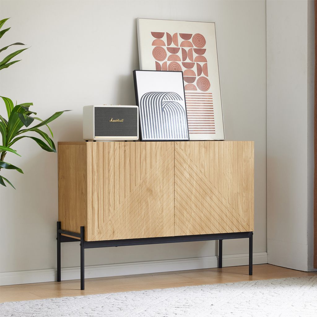 A wooden sideboard with black metal legs, decorated with two framed art prints and a speaker, placed on a rug in a room.