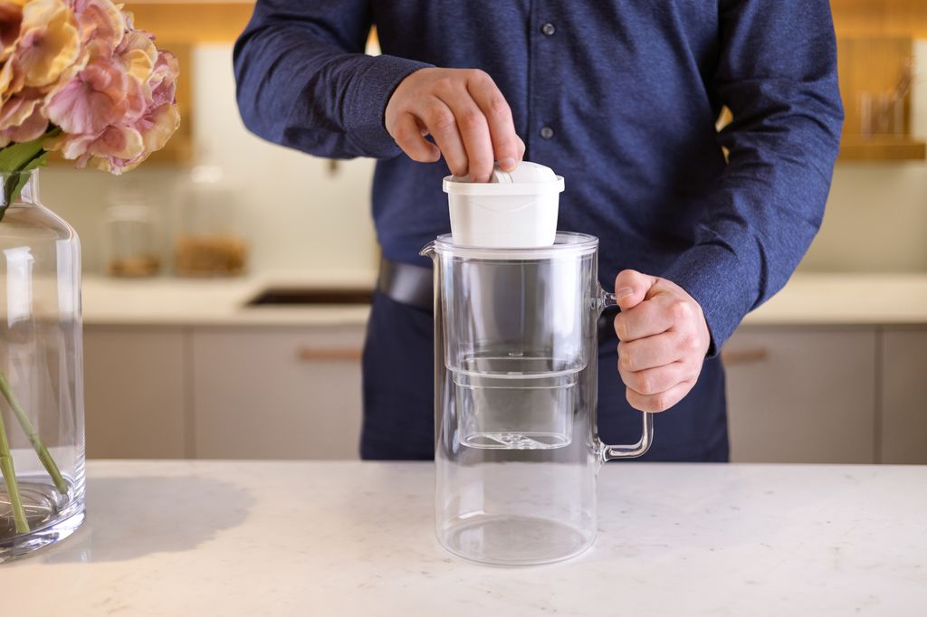 A person is placing a filter into a glass water pitcher on a countertop.