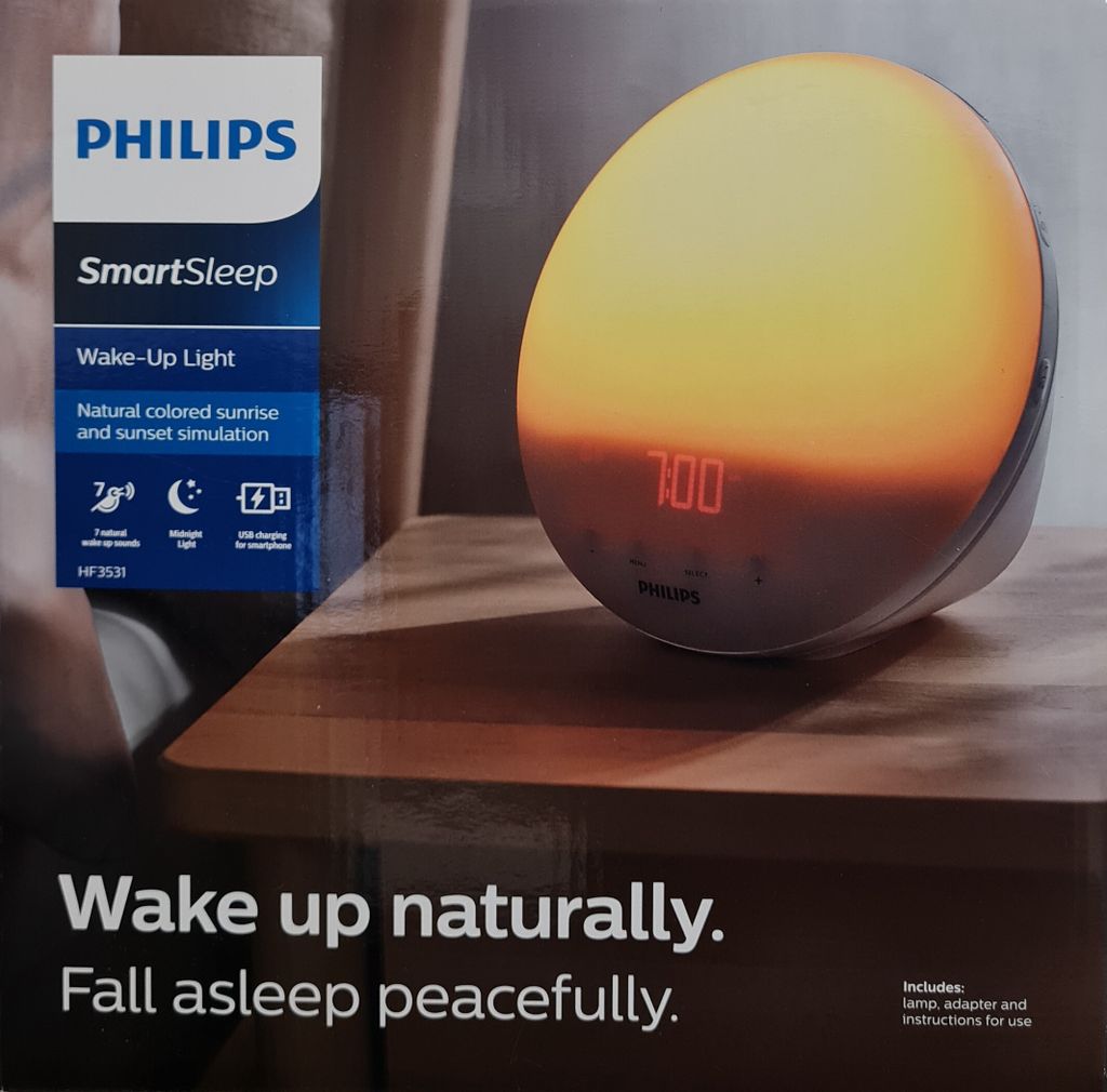 A Philips SmartSleep Wake-Up Light is displayed on a wooden table, simulating a sunrise.