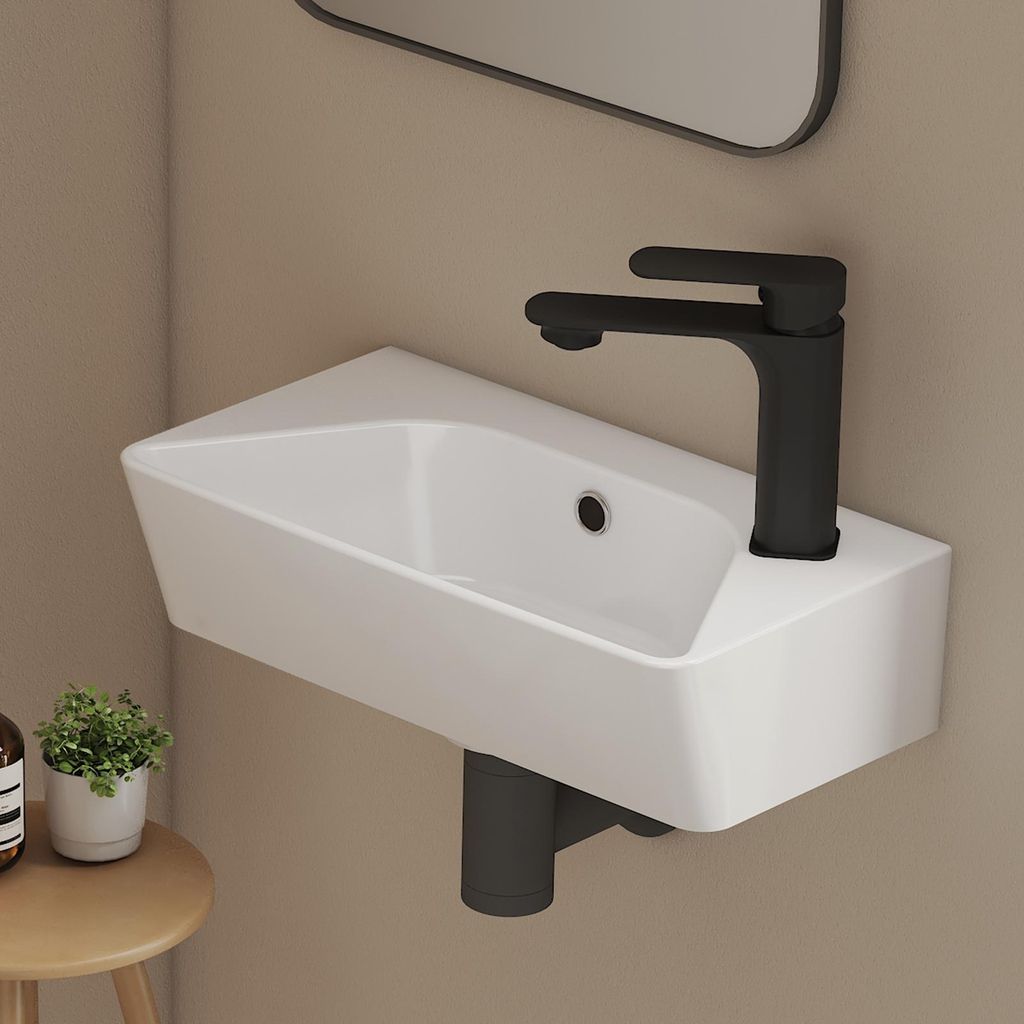 A modern white ceramic wall-mounted sink with a black faucet is shown against a light beige wall. A small potted plant and a bottle are visible on a wooden stool to the left.