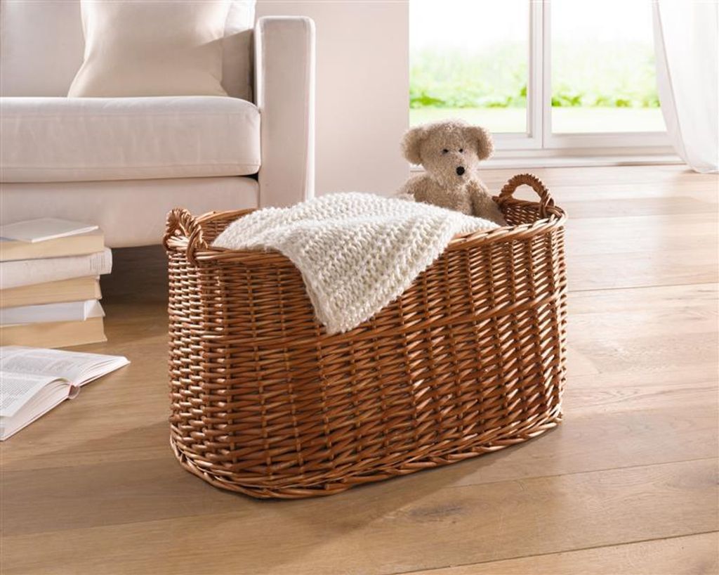 A wicker basket with a blanket and a teddy bear sits on a wooden floor next to a sofa. A window with greenery is visible in the background.
