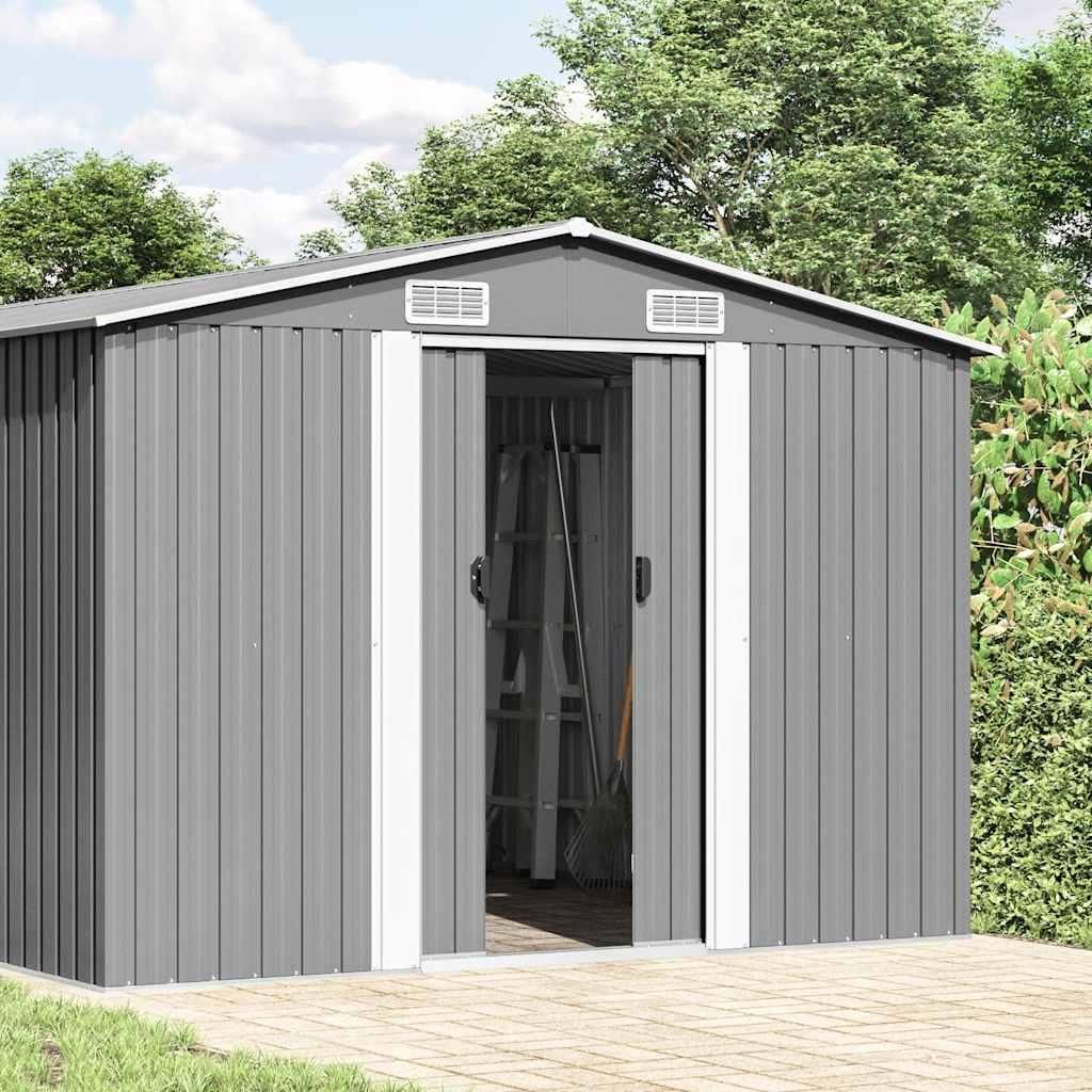 A grey metal garden shed with its doors open, showing a ladder inside. It is set in an outdoor environment with trees and a paved patio.