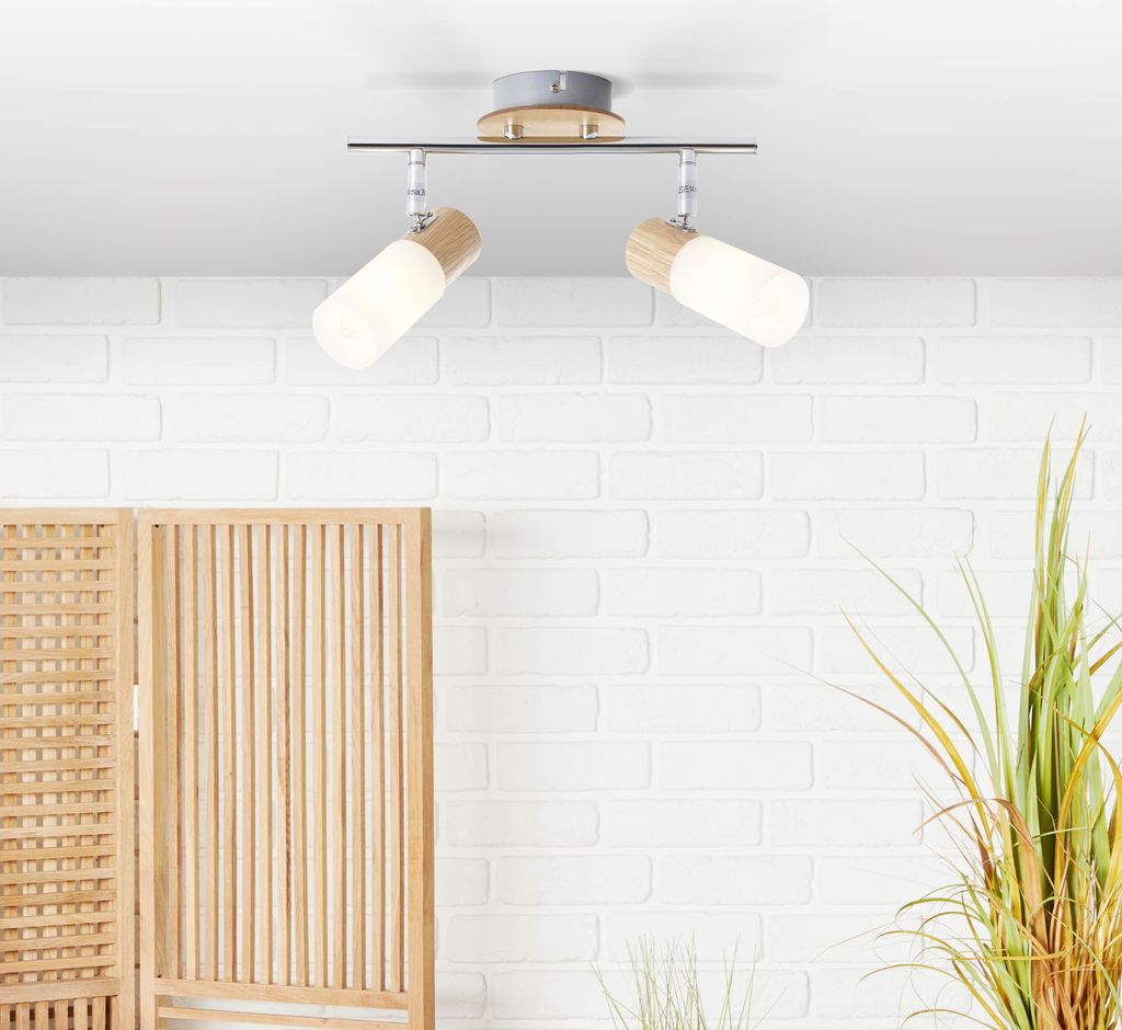 A ceiling light fixture with two adjustable spotlights is shown against a white brick wall background. A wooden room divider is partially visible on the left, and a tall green plant is on the right.