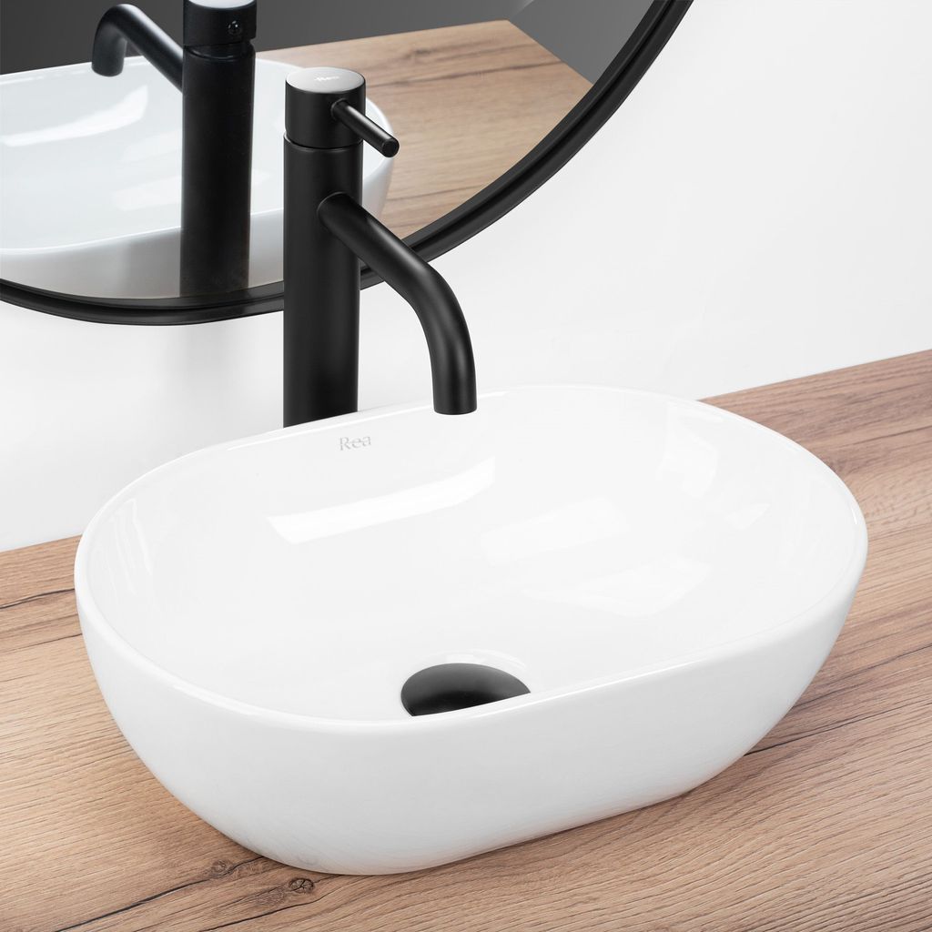 A white oval basin sink with a black faucet is shown on a wooden countertop, with a reflection in a mirror behind it.