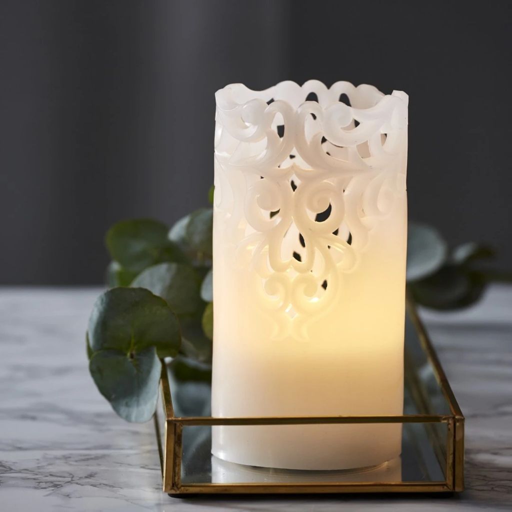 A white LED candle with a decorative filigree design sits on a mirrored tray with a brass frame, next to some green foliage.