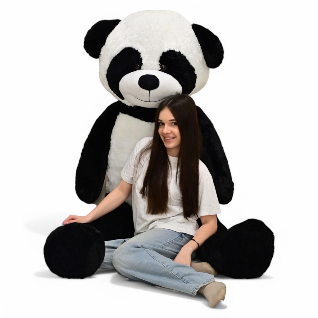 A young woman is sitting in front of a very large stuffed panda toy.
