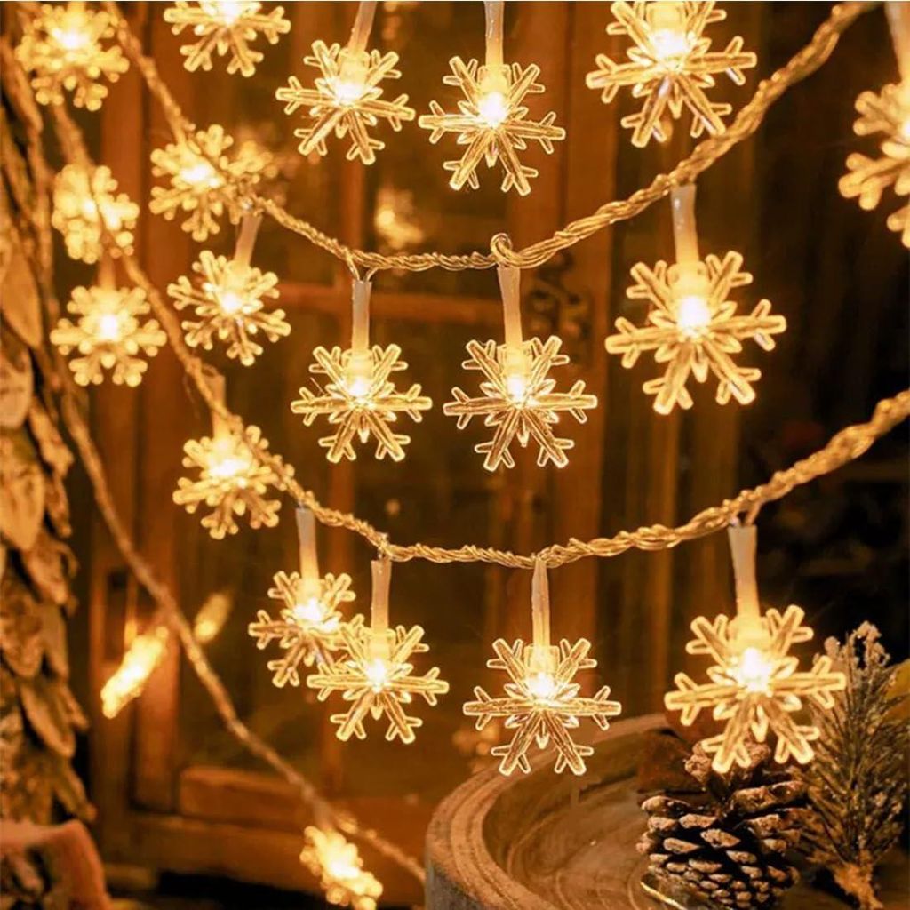 A string of snowflake-shaped lights is draped around a window or doorway, with a pinecone and greenery in the foreground.