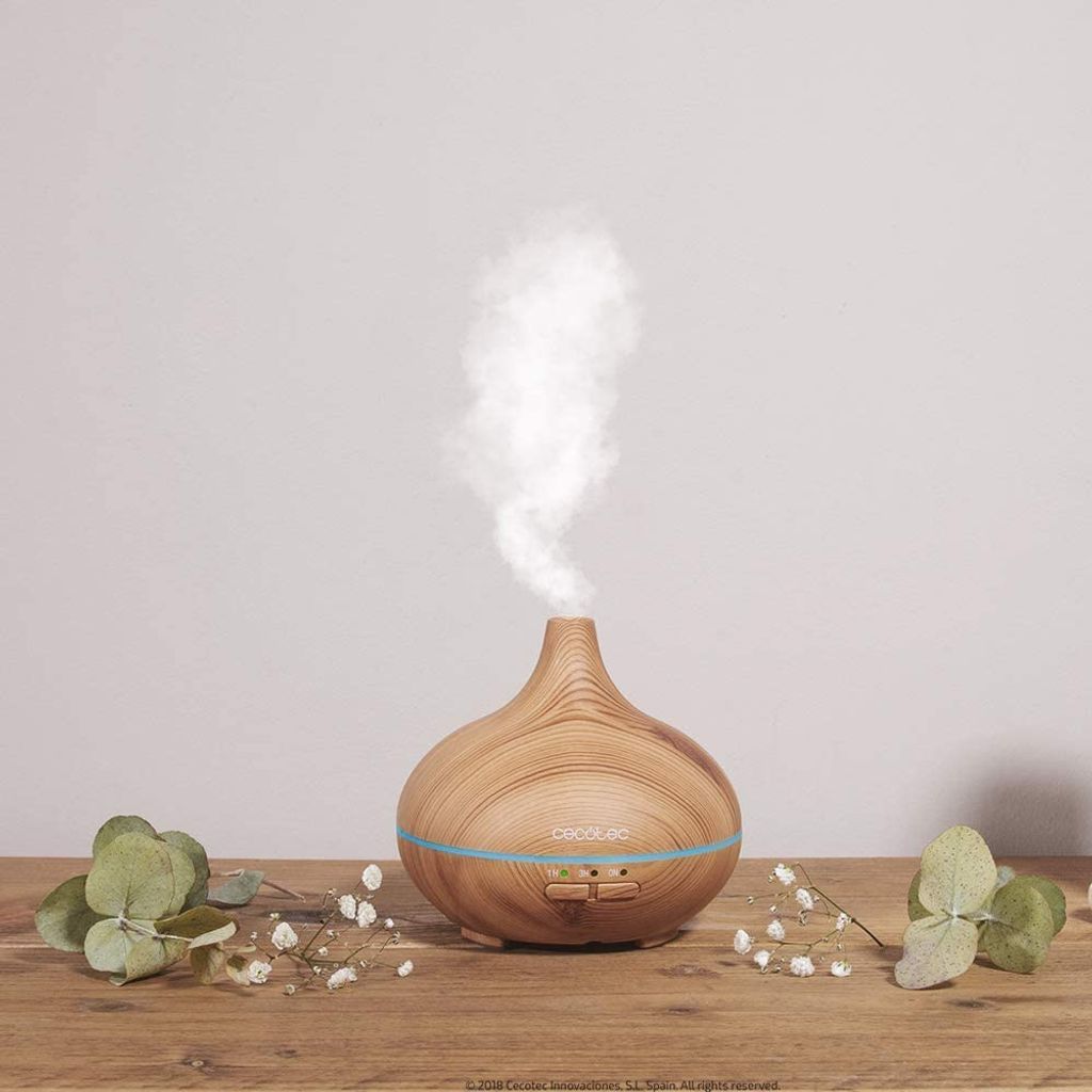 A wooden essential oil diffuser emitting mist, placed on a wooden surface with greenery and small white flowers around it.