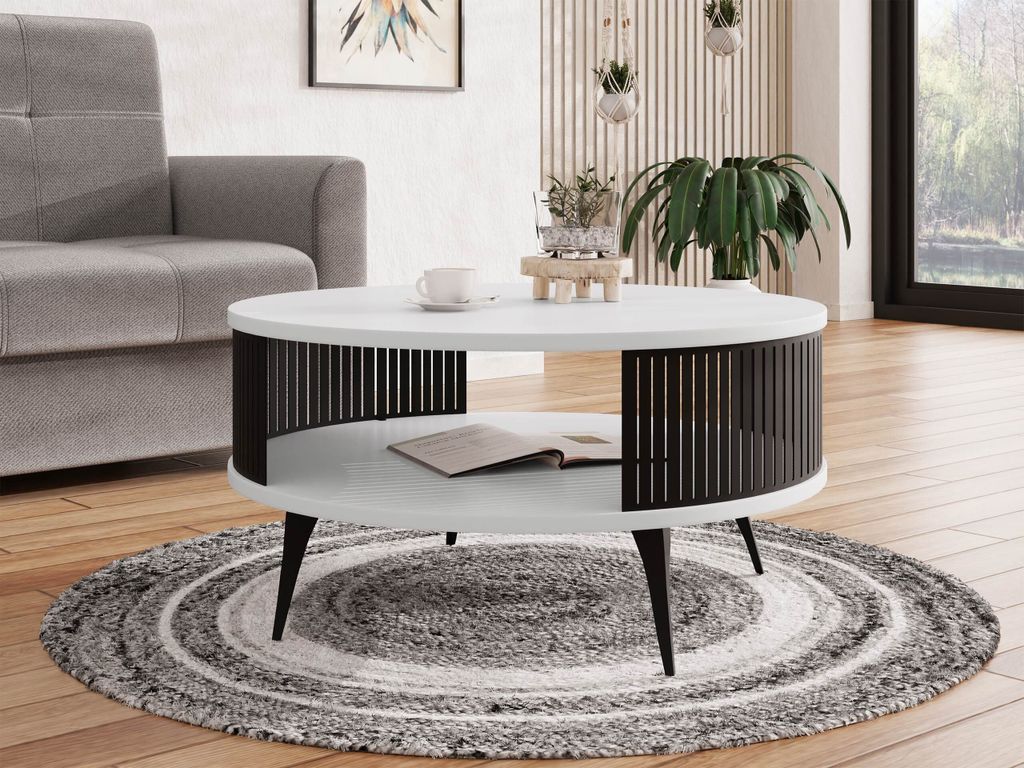 A modern living room scene featuring a round coffee table with a slatted base, placed on a circular rug. A sofa is visible to the left, and a window with greenery is in the background.
