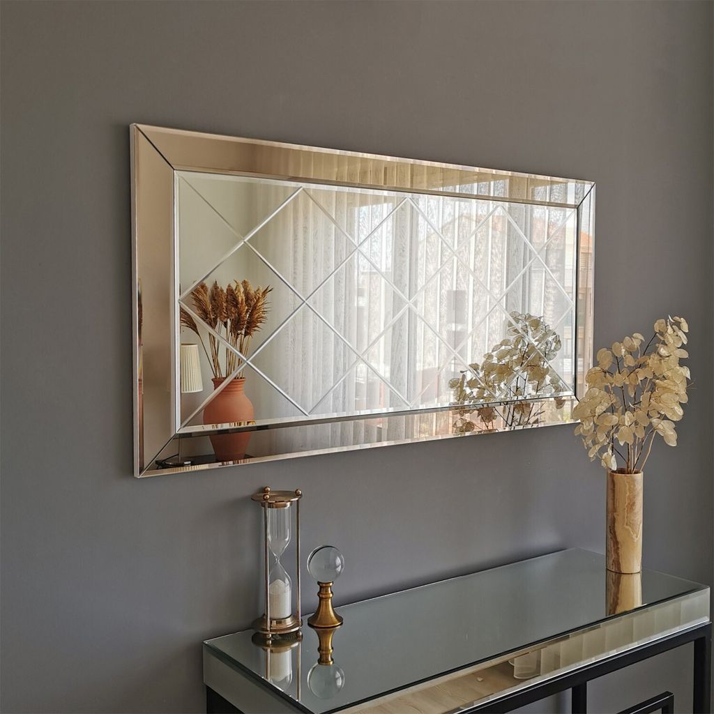 A rectangular mirror with a decorative frame hangs on a gray wall above a mirrored console table. The table holds an hourglass and a vase with dried flowers. Another vase with dried flowers is reflected in the mirror.