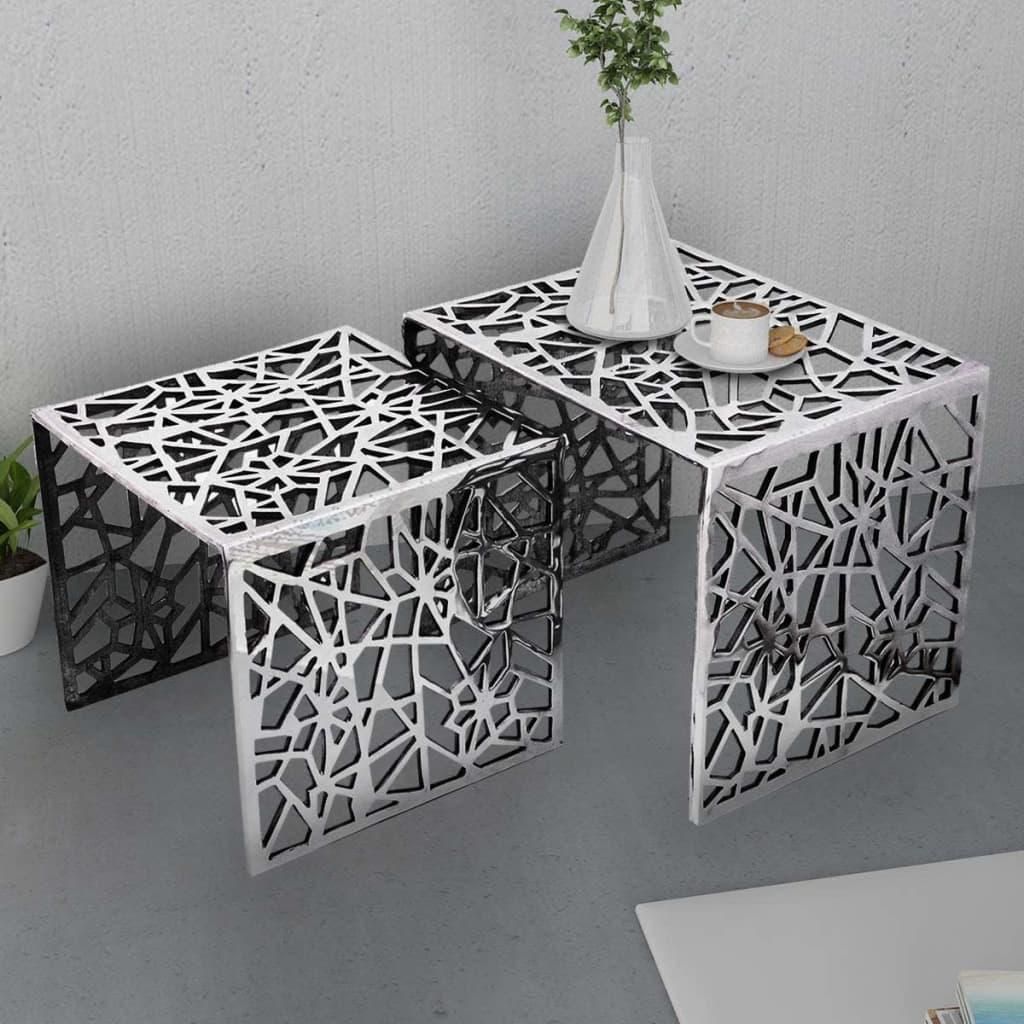 Two nesting side tables with a geometric cut-out pattern are shown in a modern interior setting. One table has a cup of coffee and a cookie on it, and a vase with greenery is placed on the larger table.