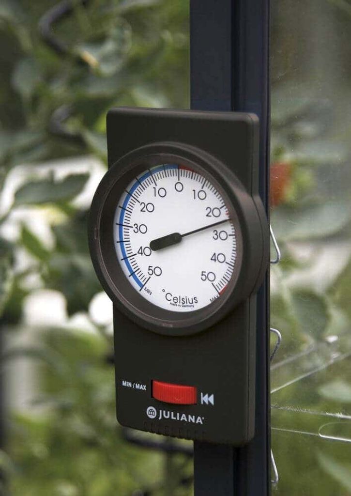 A thermometer with a black casing and a white dial is mounted on a dark pole, with a blurred green background of plants.