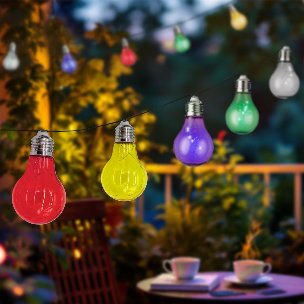 A string of colorful LED light bulbs hanging outdoors at night, with a table and chairs in the foreground.