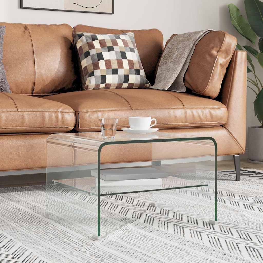 A glass coffee table is placed in front of a brown leather couch, with a cup of coffee and a glass of water on it.
