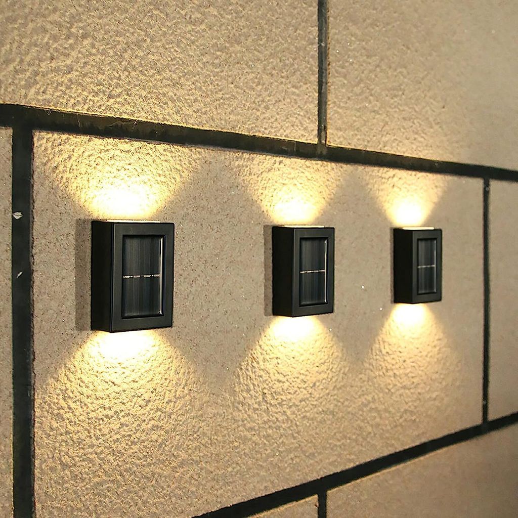 Three black solar wall lights are mounted on a textured, light-colored wall with dark grout lines. The lights are illuminated, casting warm yellow light upwards and downwards.