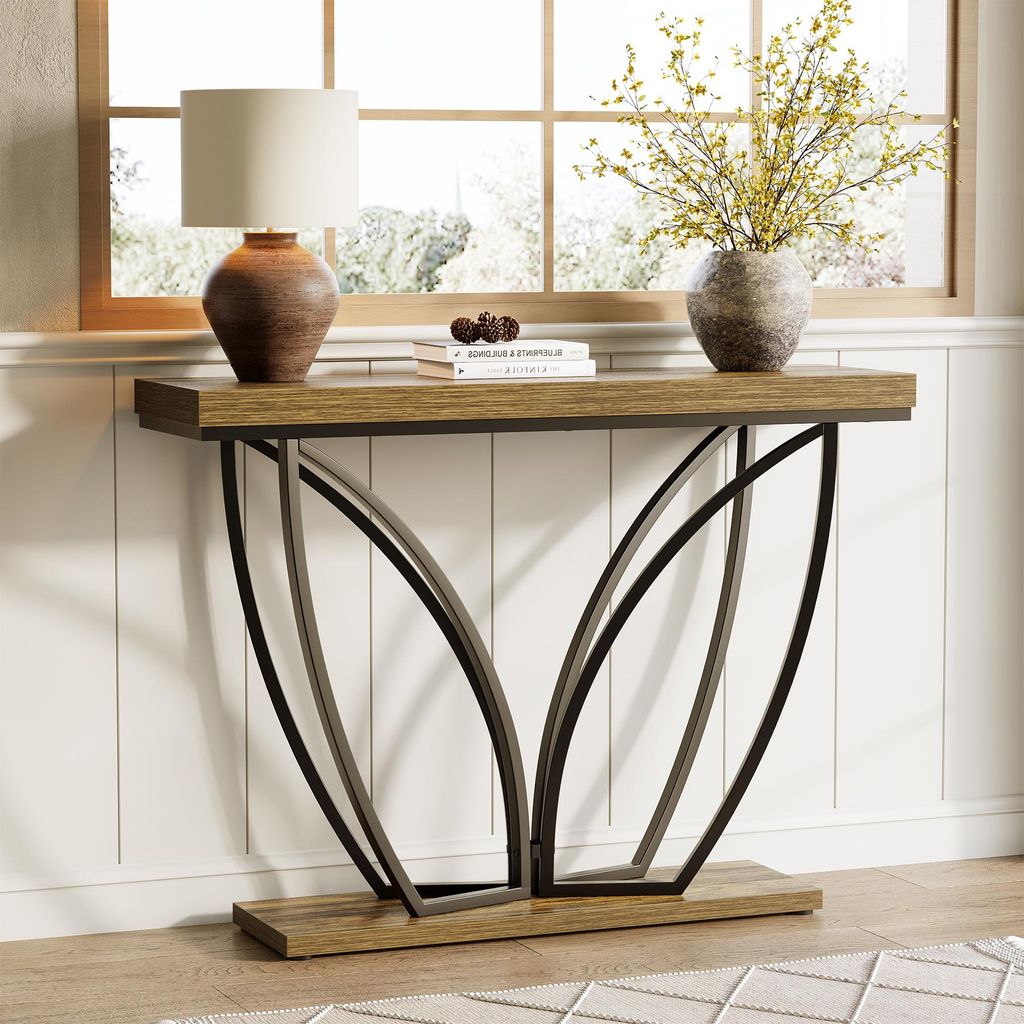 A console table with a lamp and vase of flowers on it, placed against a white paneled wall and a window.