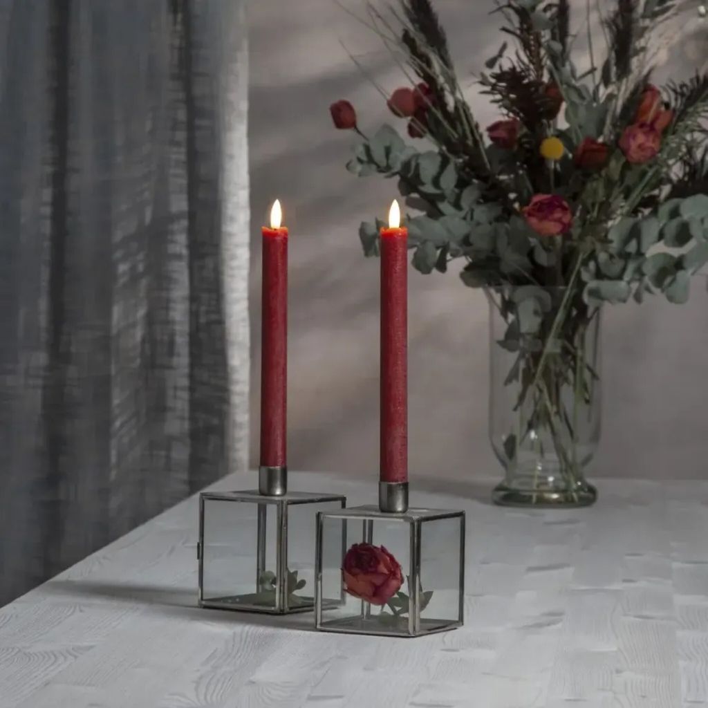 Two red LED taper candles in glass candle holders on a white table, with a floral arrangement in the background.
