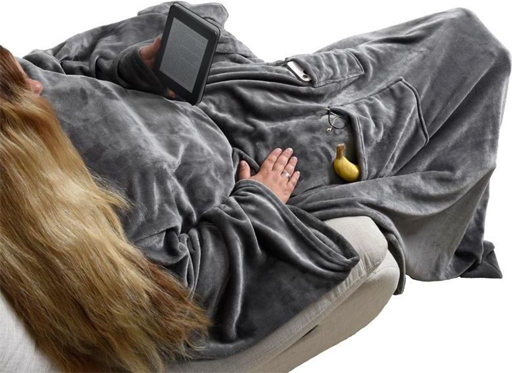 A person is lying on a couch, wrapped in a large gray blanket with sleeves, holding an e-reader.