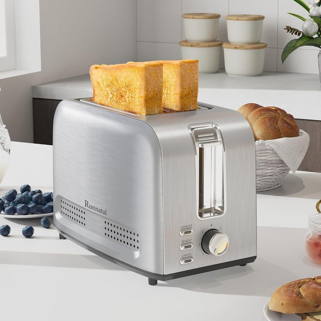 A silver toaster with two slices of toast in it, placed on a white countertop with a bowl of blueberries and some bread rolls in a basket in the background.