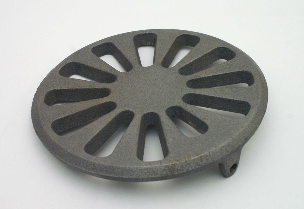 A round, cast iron grate with a spoke-like pattern and a small protrusion on the side, set against a white background.