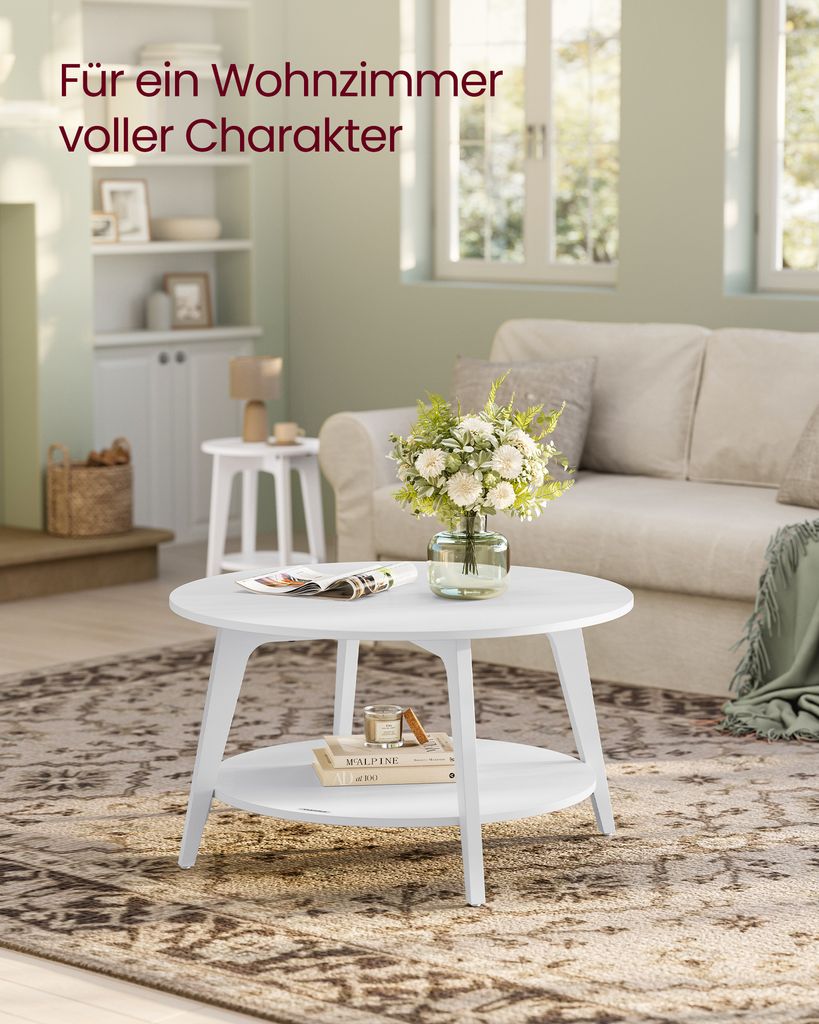 A white round coffee table with a shelf is placed on a patterned rug in a living room. A vase of flowers sits on the table, and a sofa is visible in the background.