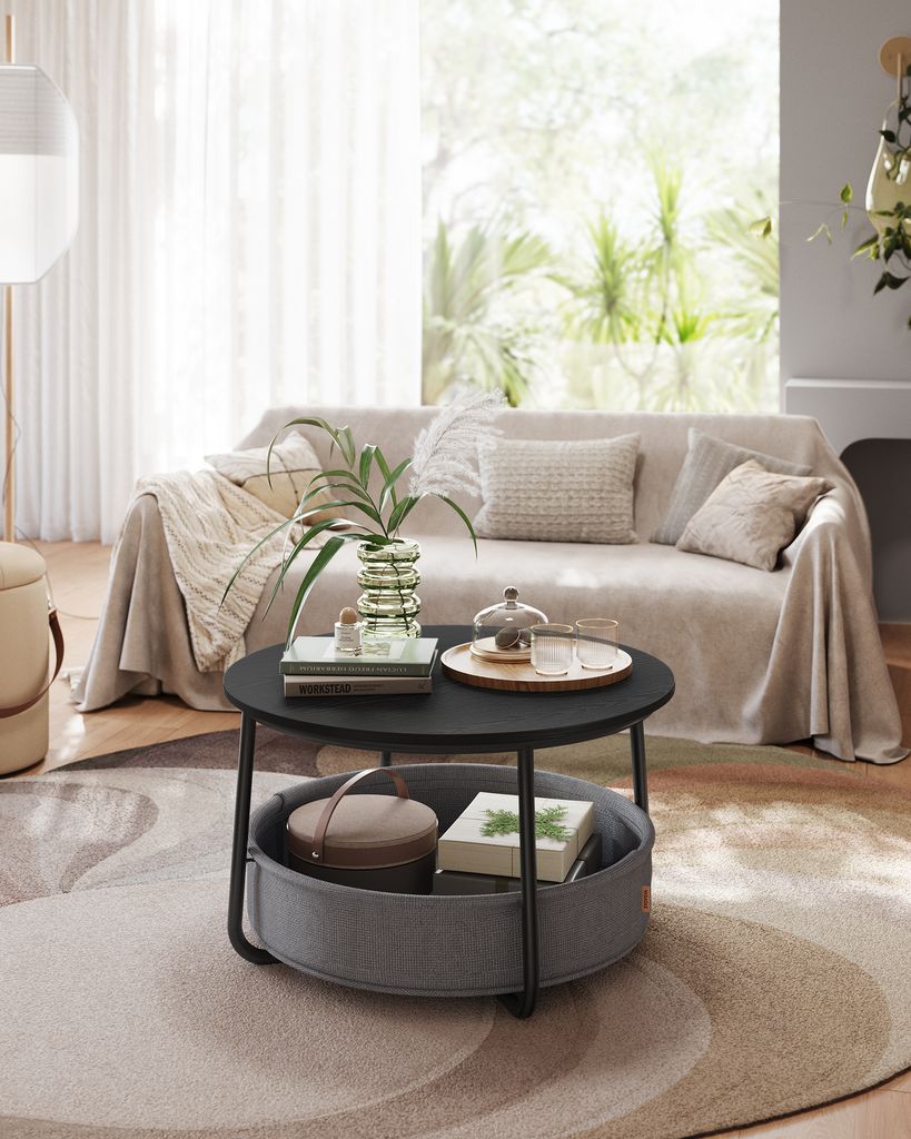 A living room scene featuring a round coffee table with a fabric basket underneath, placed in front of a sofa. The scene is well-lit and styled.