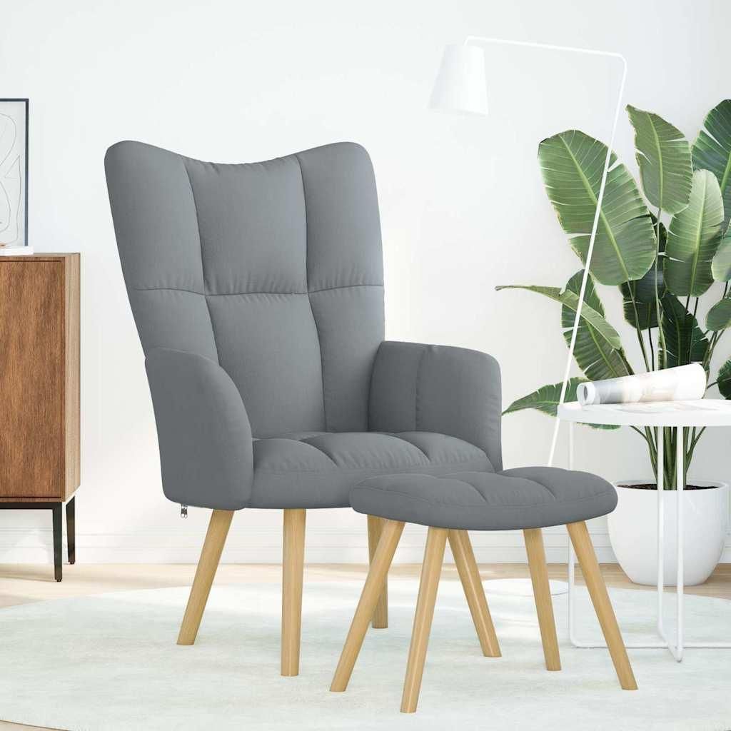 A gray fabric armchair and ottoman are shown in a well-lit room with a large plant and a floor lamp.