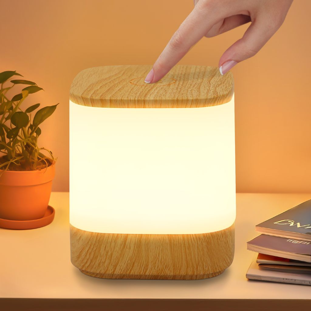 A hand is touching a wooden-topped LED lamp on a white surface, with a potted plant to the left and books to the right.