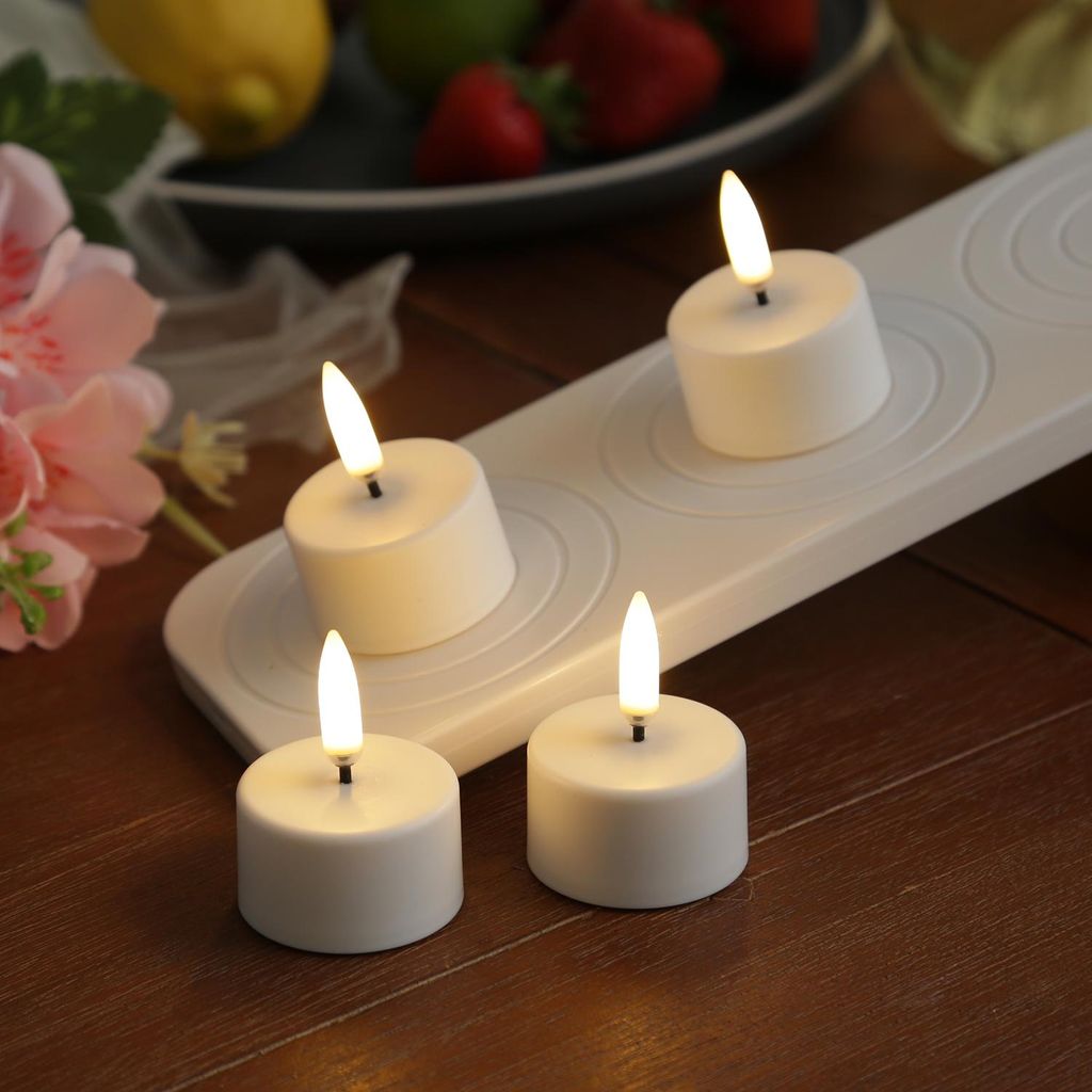 Four LED tea lights with a charging station are arranged on a wooden surface, with some flowers and fruit in the background.