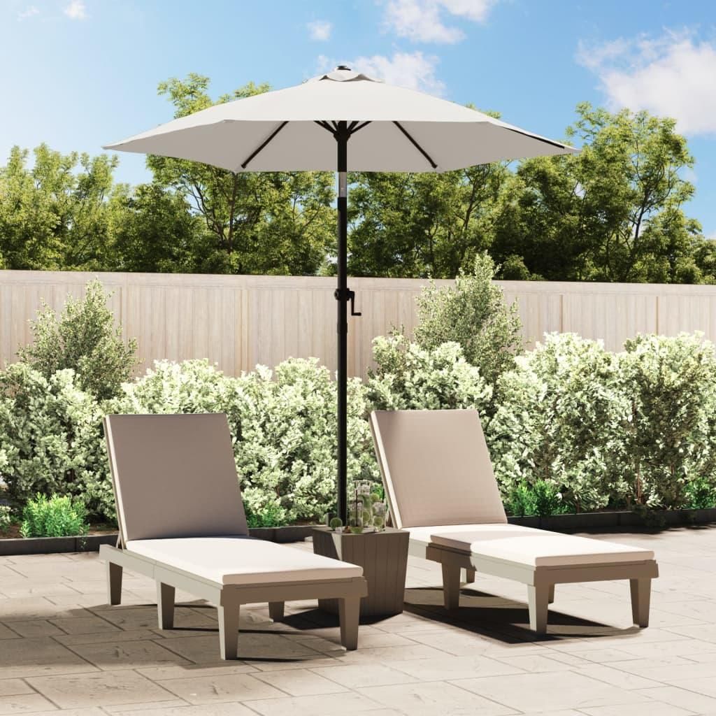 Two lounge chairs and a patio umbrella are set up on a paved patio area with lush green bushes and a wooden fence in the background.