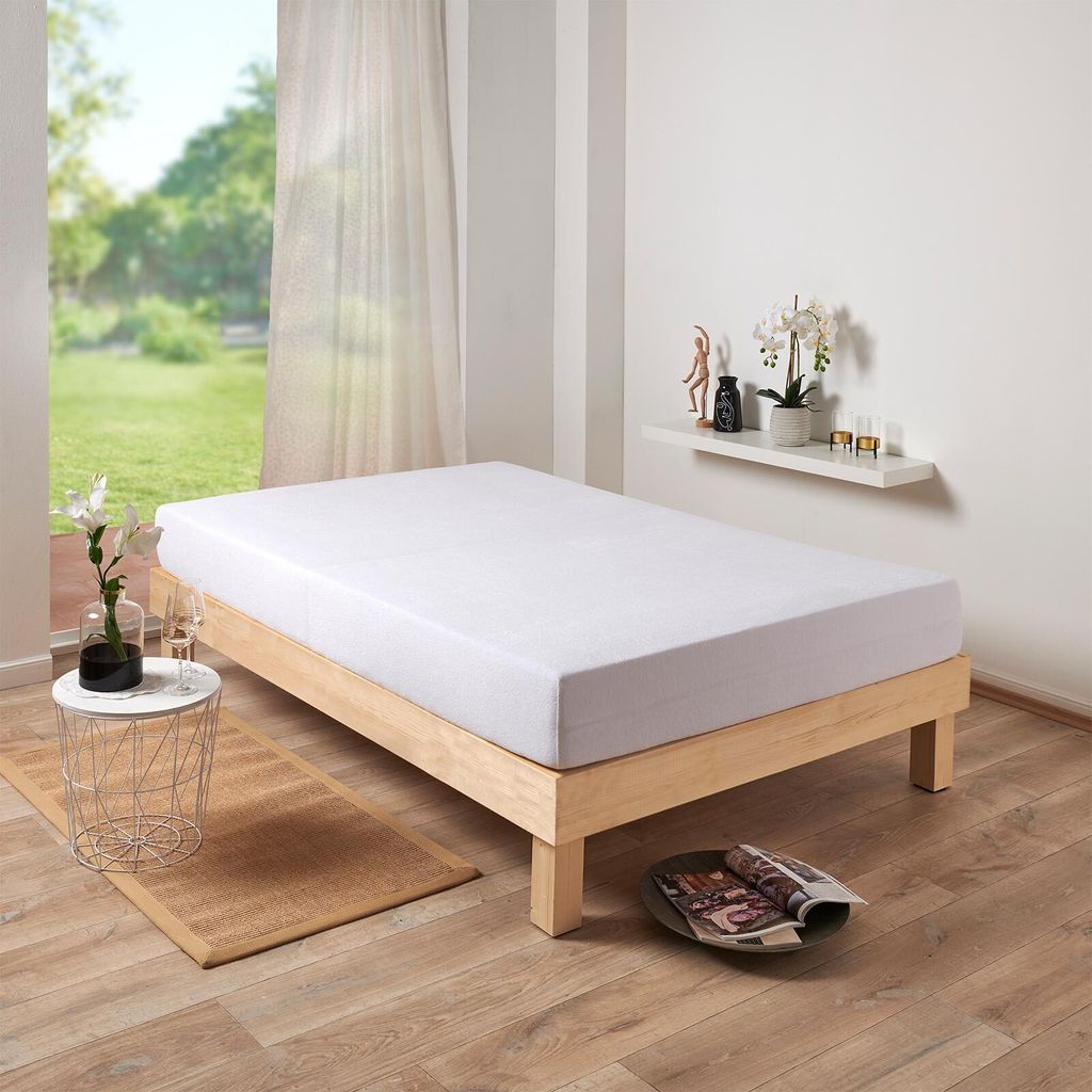 A white mattress on a wooden bed frame, in a room with a window and some decorative items on a shelf.