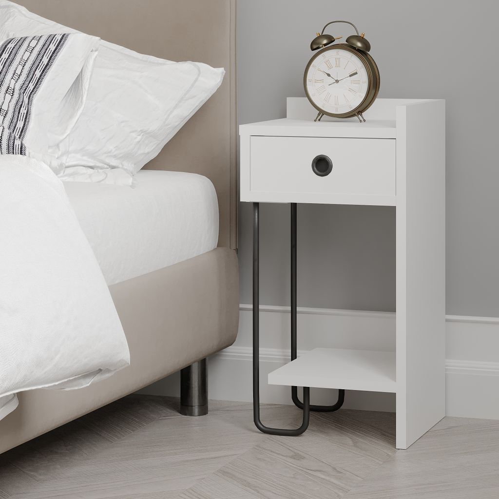 A white bedside table with a drawer and a shelf, next to a bed. An alarm clock sits on top of the table.