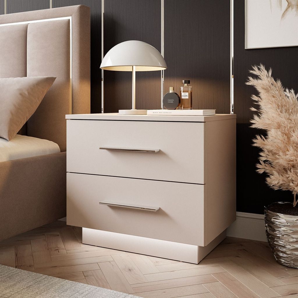 A modern bedside table with two drawers and LED lighting is shown next to a bed in a bedroom setting. A lamp and two perfume bottles are on top of the table.