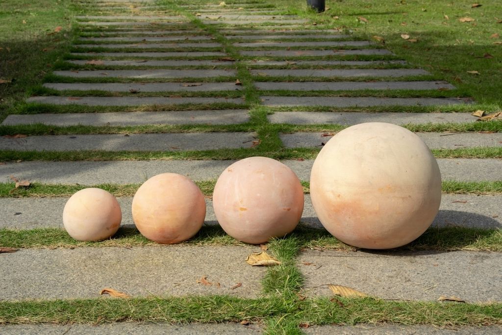 Four terracotta spheres of varying sizes are placed on a pathway with grass growing between the stones.