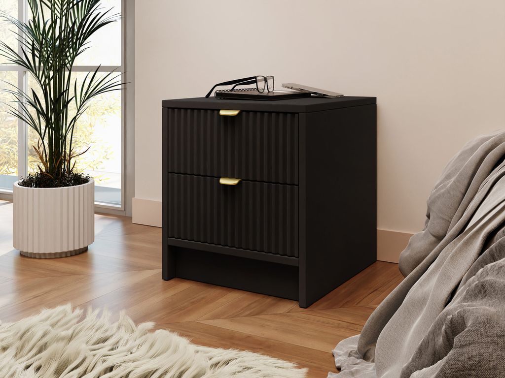 A black nightstand with two drawers and gold handles is placed next to a bed. A plant is in a white pot to the left of the nightstand, and a rug is in the foreground.