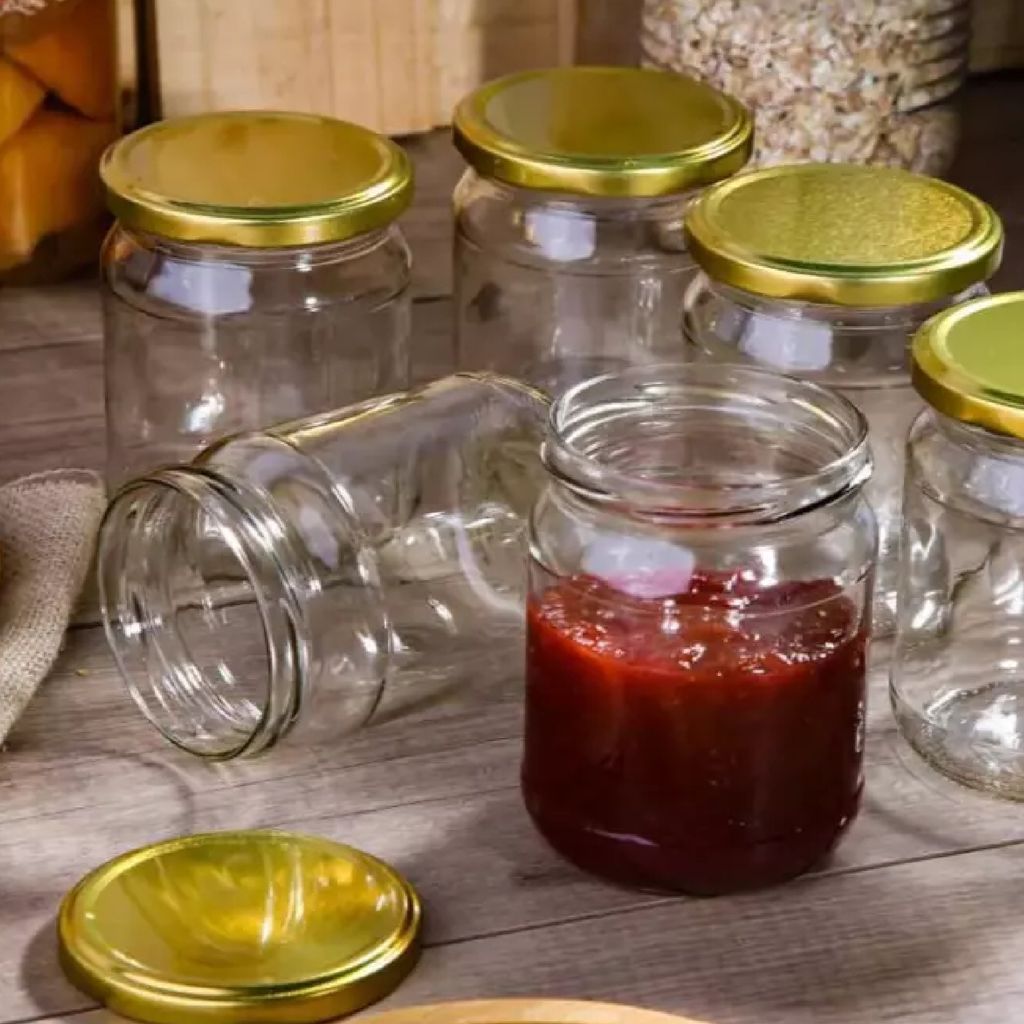 Several glass jars with gold lids are arranged on a wooden surface. One jar in the foreground contains a red jam or preserve.